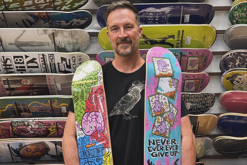 Brady Smith holding the two Neuro decks he signed, standing in front of a wall of skateboards.