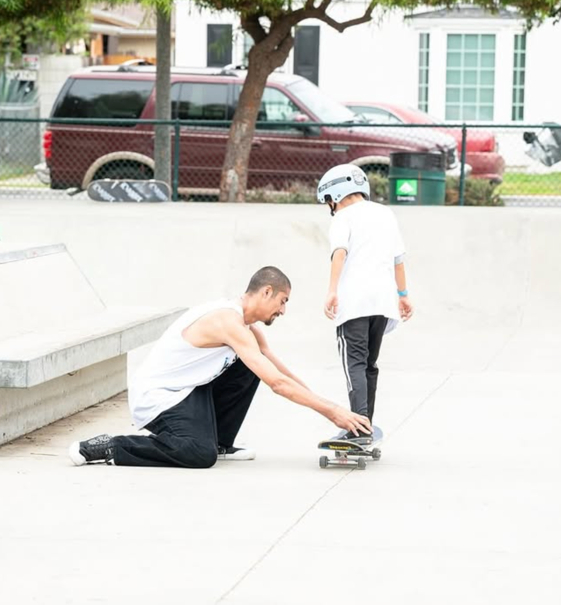 Coach Vincent helps a young skater in a helmet balance on a skateboard at a skatepark.
