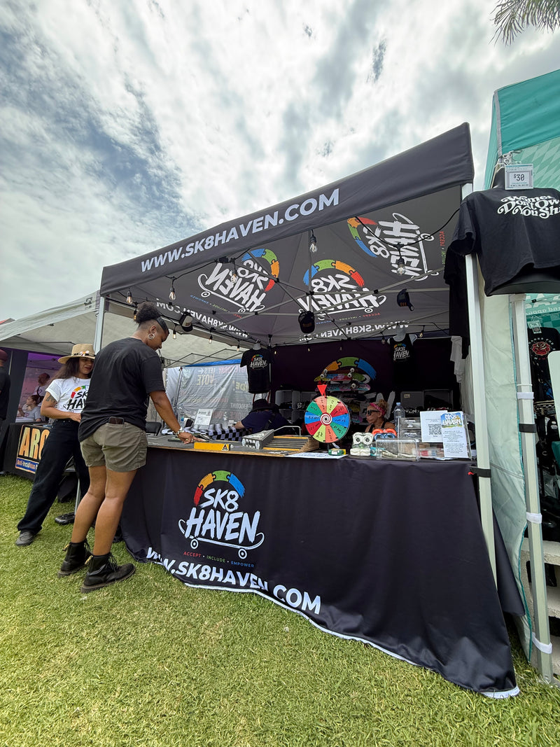 The Sk8 Haven booth at Warped Tour, a black canopy printed with the Sk8 Haven logo, a prize wheel on the counter and staff talking with visitors.