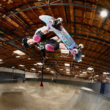 A skateboarder in a black helmet and pink knee pads performs a trick mid-air above a concrete skate bowl inside a large indoor arena with a wooden ceiling.