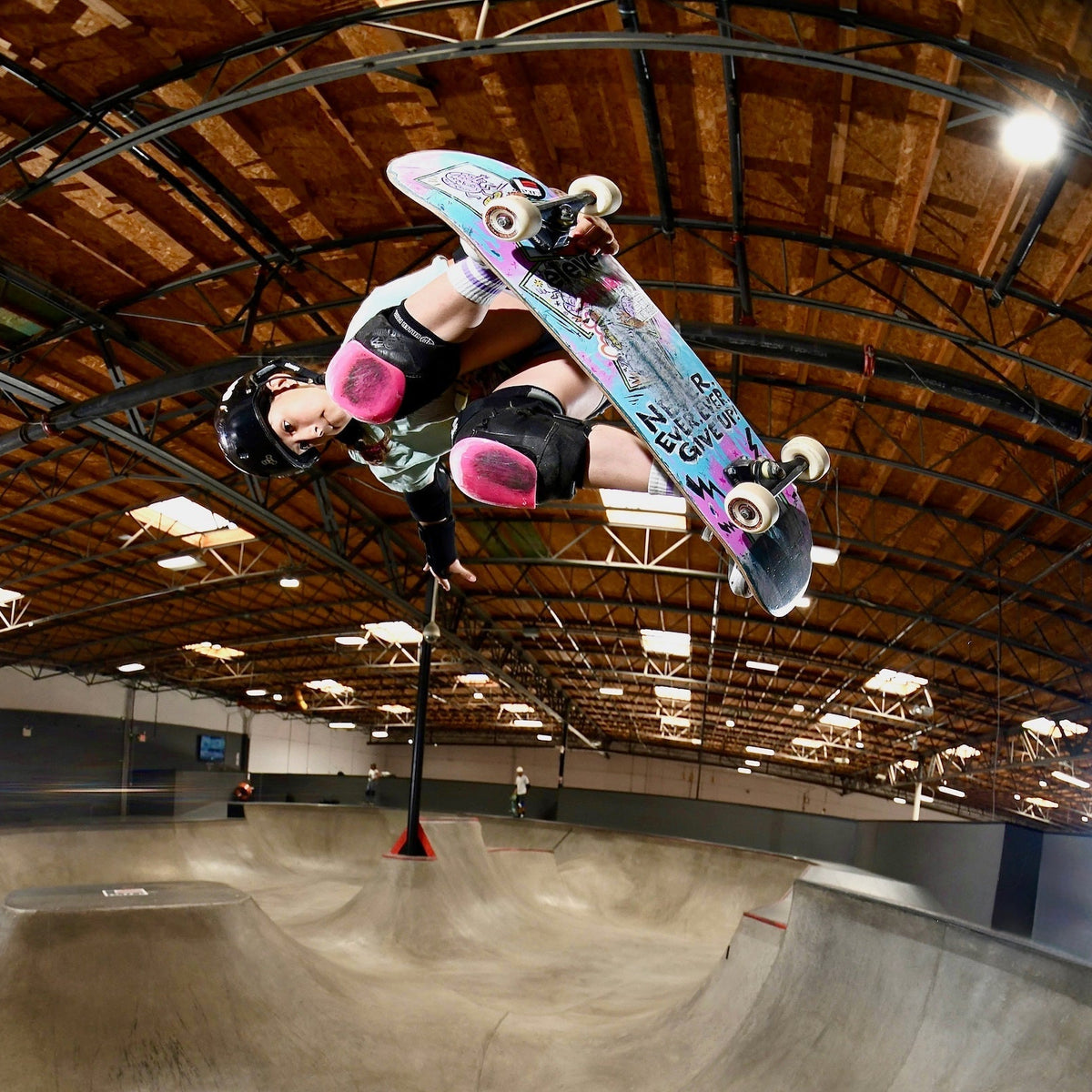 A skateboarder in a black helmet and pink knee pads performs a trick mid-air above a concrete skate bowl inside a large indoor arena with a wooden ceiling.