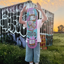 A skater tosses a Never Give Up deck into the air in front of a graffiti-covered shipping container at sunset, the board hanging in front of them mid-throw.