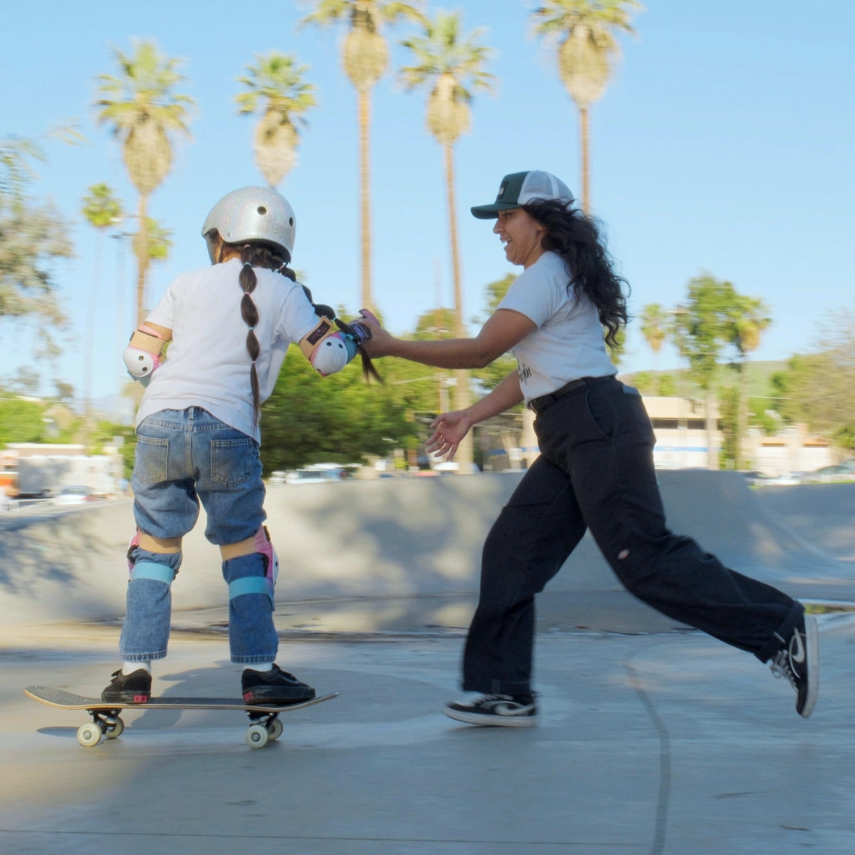 Coach Yvette from Skate Inclusion runs next to a little girl on a skateboard in the flat ground of a skate park after the little girl dropped in from a quarter pipe. Palm trees and a blue sky in the background.